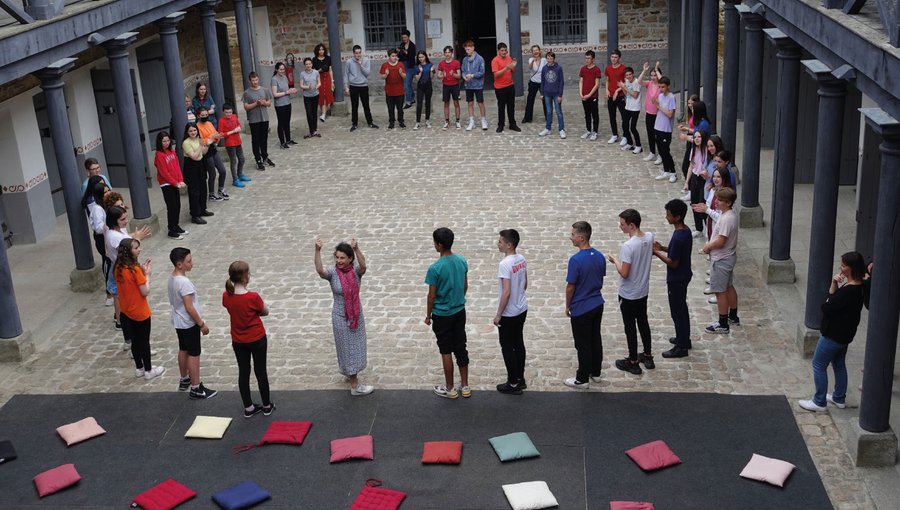 Sur cette image, un groupe d'une trentaine de personnes se tient en cercle dans la cour pavée de la prison de Guingamp. Une animatrice au centre, habillée en blanc avec une écharpe rose, semble guider les participants. Les personnes, majoritairement des jeunes, portent des vêtements de couleurs variées et sont attentives à l'animation en cours. Des coussins sont disposés au sol sur un tapis gris à l'extérieur du cercle.